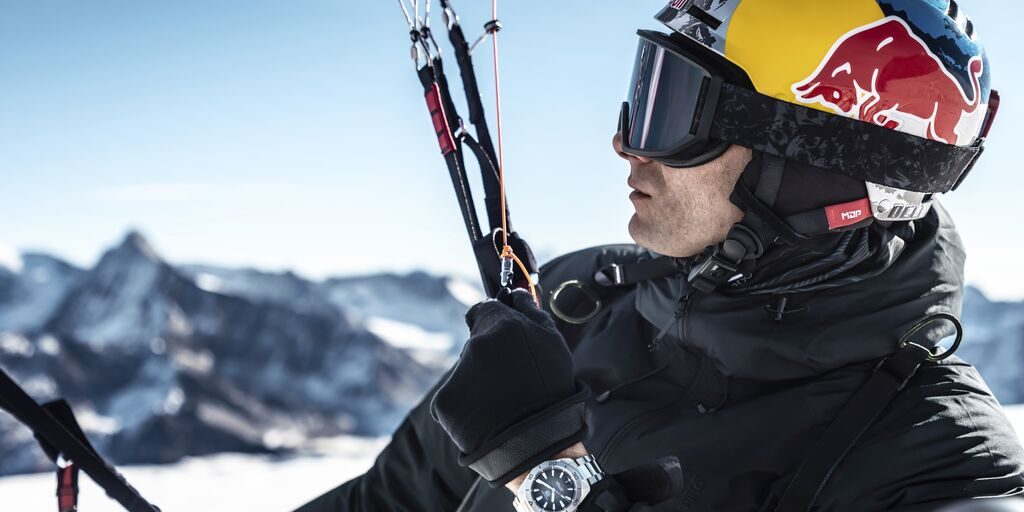 Man with snowy mountain background wearing a team red bull helmet, with black coat and black dial Aquaracer watch.