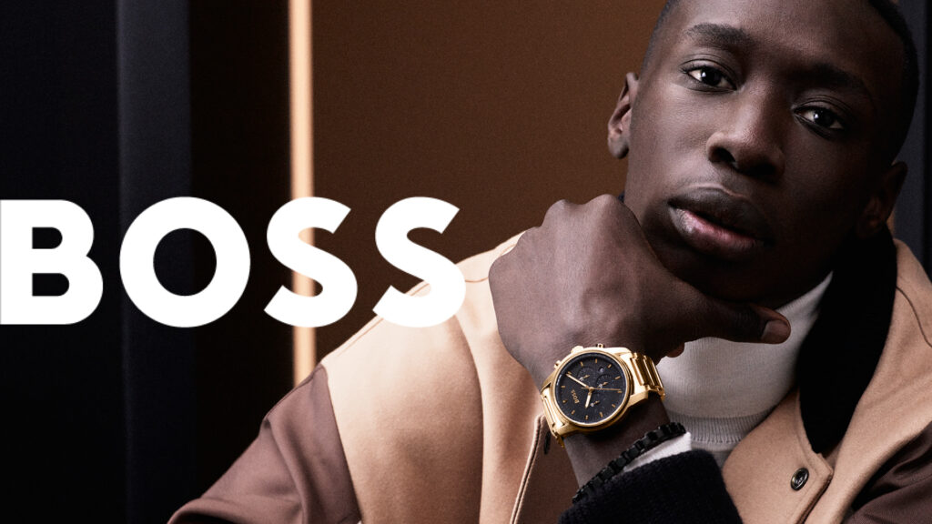 Model in brown jacket and white jumper wearing a gold BOSS watch 