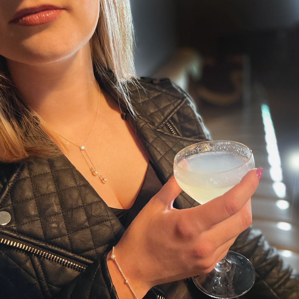 Lady wearing a black leather jacket with a matching silver and pearl necklace and bracelet set, holding a cocktail in her hand