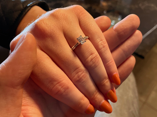 a ladies hand with dark orange nails with a emerald cut diamond ring in platinum on her engagement finger. 